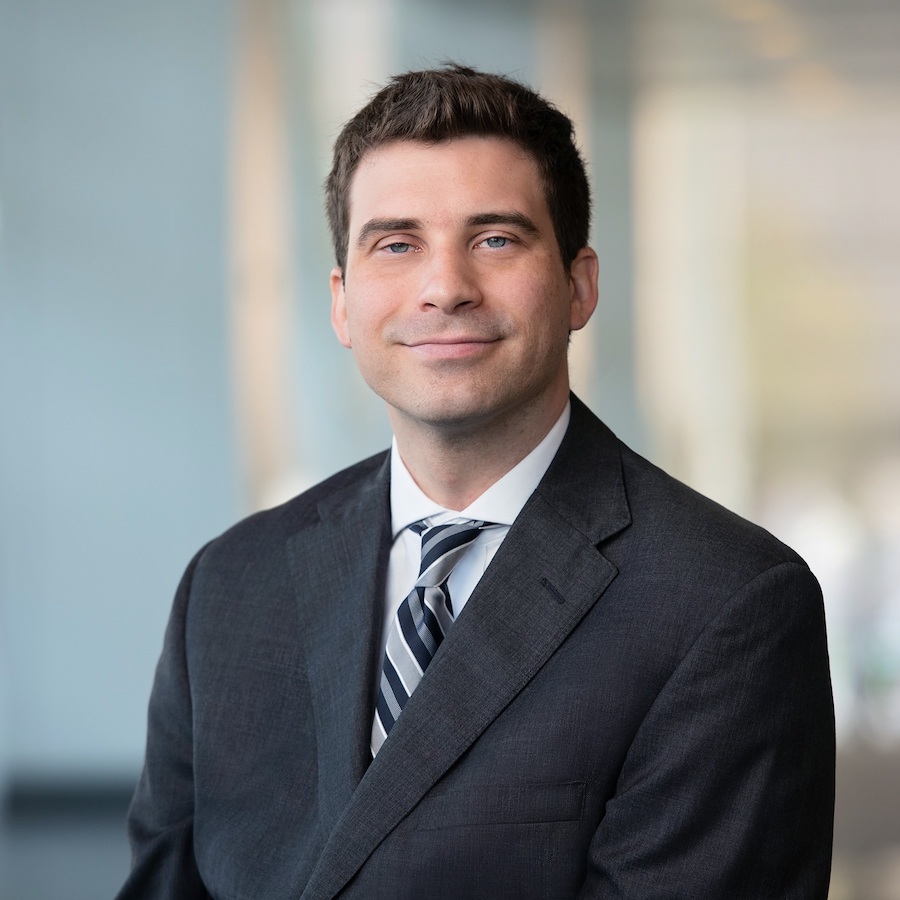 Headshot of a smiling man in a dark grey suit and a striped blue and white tie.