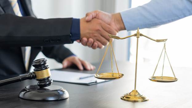 A lawyer shakes their client's hand. On his desk is paperwork, a gavel, and the scales of justice.